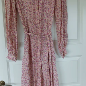 Blommig långklänning med skärp i midjan,  - Blommig långklänning från 2Day.  Klänningen är gul, rosa och blå, den har skärp i midjan och knappar fram hela vägen.  Den köptes nyligen second hand på Arkivet och jag skulle säga att den är i bra skick. 