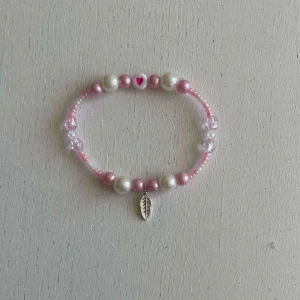Egengjort pärlarmband  - Ett egengjort pärlarmband med färgerna rosa och vit i olika nyanser med en blandning pärlsorter. Armbandet har en berlock i silver som avbildar en fjäder. 