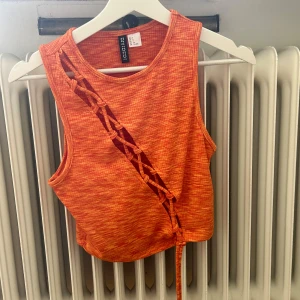 Orange ärmlös topp från Bershka - En trendig orange ärmlös topp från HM med ett unikt snörningsmönster på framsidan. Toppen har en rund halsringning och en knytning i nederkanten.