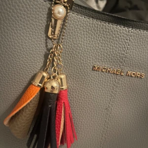 Oanvänd Michael Kors väska - Grå väska med fina detaljer bla pärlor vid fästen och ”tofsar” i rött, svart, brun/orange. Höjd 22cm. Bredd 30cm. Axelrem medföljer. 