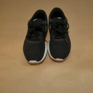 Svarta Nike sneakers - Ett par svarta Nike sneakers med vit sula och vit Nike-logga på sidan. Skorna har snörning och är i använt skick.