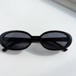 Svarta ovala solglasögon - Ett par svarta ovala solglasögon med mörka linser. Glasögonen har en enkel och stilren design med en svart ram och svarta skalmar.