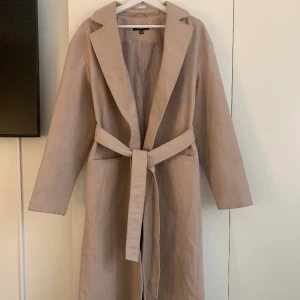 Lång kappa - En lång beige kappa från Missguided i storlek 40 (passar mer som en 38 enligt mig). Jättefint skick och passa för vår/höst. Fickor framtill samt band man knyter i midjan. 