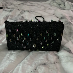 Svart glittrig kuvertväska - Säljer en superfin svart kuvertväska med glittriga detaljer. Väskan har en axelrem och är perfekt för festliga tillfällen. Den är dekorerad med små pärlor och paljetter som ger en extra touch av glamour. Väskan har en dragkedja för säker förvaring av dina saker.