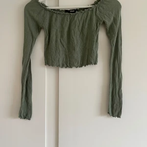 Grön ribbad topp från Bik Bok - Säljer en superfin grön ribbad topp från Bik Bok i storlek XS. Toppen har långa ärmar och en croppad passform med volangdetaljer vid halsringningen, ärmsluten och nederkanten. Perfekt för både vardag och fest! 💚