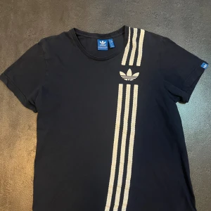 Marinblå t-shirt från Adidas - Säljer en mörkblå t-shirt från Adidas med deras klassiska logga och tre ränder i vitt på framsidan. T-shirten är kortärmad och gjord i bomull, vilket gör den superbekväm att ha på sig. Perfekt för både vardag och träning!