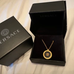 Guldhalsband från Versace - Säljer ett snyggt guldhalsband från Versace. Halsbandet har en rund medaljong med Versaces ikoniska Medusa-huvud i mitten och en grekisk nyckel-design runt kanten. Perfekt för att ge din outfit en lyxig touch!