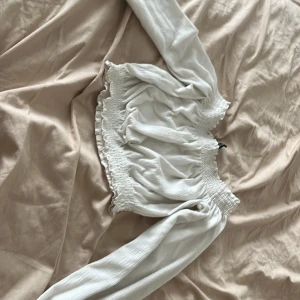 Vit ribbad topp med volangdetaljer - Säljer en superfin vit ribbad topp i storlek XS. Den har långa ärmar och en söt volangdetalj vid midjan. Perfekt för både vardag och fest! Toppen är i nyskick och väldigt bekväm att ha på sig.