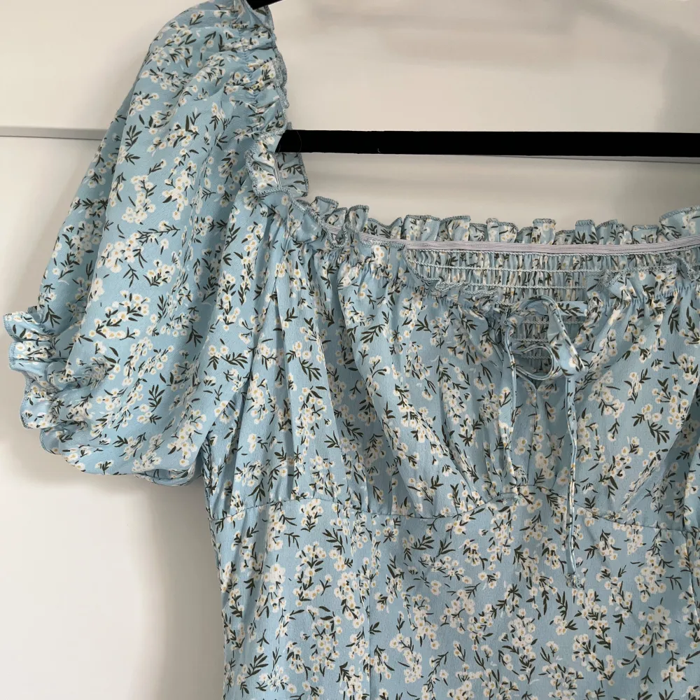 Ljusblå klänning med vita blommor, går att ha som offshoulder eller med armarna uppåt. Se detaljer på bilderna. Helt oanvänd . Mekot.