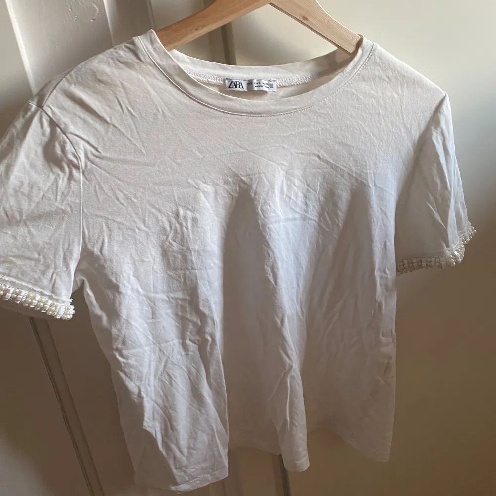 Säljer en superfin vit t-shirt från Zara i storlek S. Den har korta ärmar och är dekorerad med pärlor vid ärmsluten, vilket ger en extra touch av elegans. Perfekt för både vardag och fest! 💫. Toppar.