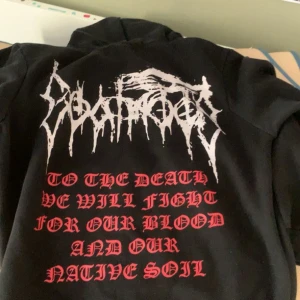 Svart hoodie med tryck från Goatmoon - Säljer en svart hoodie från Goatmoon med tryck på både fram- och baksidan. På framsidan finns en stor get-symbol inuti en röd cirkel och texten 'GOATMOON' ovanför. På baksidan står det 'TO THE DEATH WE WILL FIGHT FOR OUR BLOOD AND OUR NATIVE SOIL' i röd text. Hoodien har en dragkedja och en ficka på magen. Perfekt för höst och vinter!