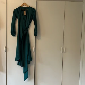 Green Satin Wrap Dress  - Klänningen har ett glansigt och silkigt material. Den är grön med blåa toner. Endast provad, bra skick förutom en nästintill osynlig rand på ryggen. Märke Enme Style produktkod på hemsidan 03377  Nypris 1131 kr