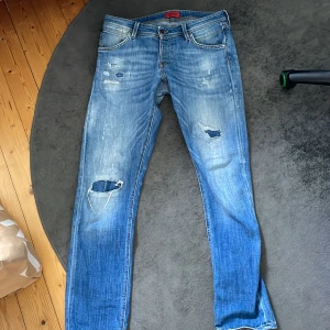 Blå slitna jeans från Jack & Jones - Säljer ett par blå jeans från Jack & Jones i storlek 34/34. De är slitna med hål och slitningar på framsidan för en cool och avslappnad look. Perfekta för vardagsbruk och ger en trendig touch till din outfit. De har en klassisk femficksdesign och en bekväm passform. Jeansen efter liknar Dondup men för en mycket mindre peng😀