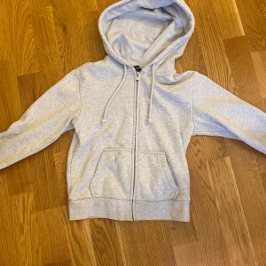 Grå hoodie från H&M - Säljer en supermysig grå hoodie från H&M i storlek XS. Den har en dragkedja framtill, två fickor och en justerbar huva med snören. Perfekt för höst och vinter när man vill hålla sig varm och bekväm. Hoodien är i bra skick och gjord av mjuk bomull. Har en liten fläck som knappt syns därför säljer jsg billigt❤️