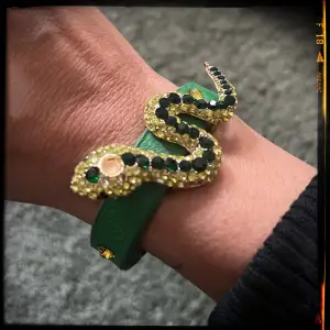 Grönt armband i skinnimmitation med orm i guld och gula stenar. Välanvänt, så lite slitage, se bild. 22,5 cm x 1,2 cm