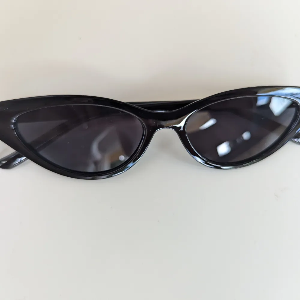 Ett par svarta solglasögon med smal båge och mörka linser. Glasögonen har en modern och stilren design som passar för olika tillfällen.. Accessoarer.