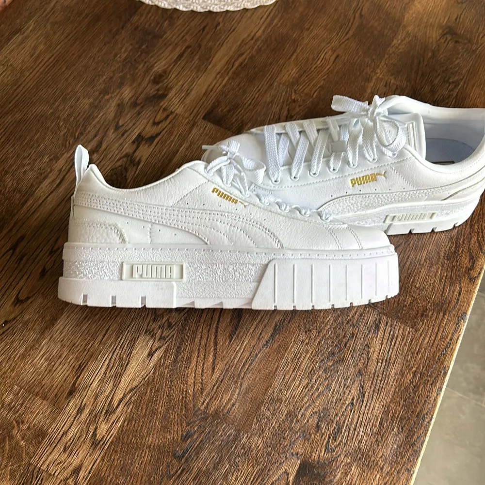 Ett par vita sneakers från Puma med snörning och en tjock sula. Skorna har en guldfärgad Puma-logga på sidan och baksidan.. Skor.