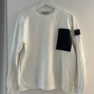 Vit tröja från Snoot - En vit långärmad tröja från Snoot med en svart ficka på bröstet och en svart logotyp på vänster ärm. Tröjan har en rund hals och är tillverkad i ett mjukt material. Tröjan är något liten i storleken. 