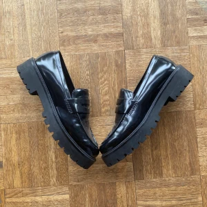 Svarta loafers - Ett par svarta loafers med en robust sula och glansig yta. Skorna har en klassisk design med en låg klack och är tillverkade i ett material som ger en blank finish.