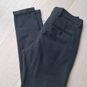Kostymbyxa Les Deux - Svarta kostymbyxor (slim-fit) från Les Deux i mycket fint skick. Storlek 30. Byxorna är stretchiga och har en smal fotöppning med uppvikt kant. En myntficka och två snedställda fickor fram samt två passpoalfickor bak. Hällor i midjan och gylf med dra