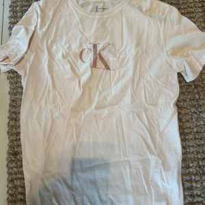 Vit t-shirt från Calvin Klein Jeans - En ljusrosa / beige t-shirt från Calvin Klein, helt oanvänd, med korta ärmar och en tryckt logotyp på framsidan. T-shirten är tillverkad av ekologisk bomull och är en del av deras hållbara kollektion.