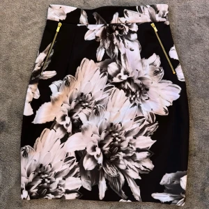 Blommig kjol med dragkedja - En elegant kjol med ett blommigt mönster i svart och vitt. Kjolen har en dragkedja både fram och bak, vilket ger en unik och stilren look. Den är knälång och har en tight passform, jättesnygg kjol, nästan ny! 
