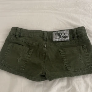 Gröna jeansshorts från Baby Phat - Ett par mörkgröna jeansshorts med slitna detaljer på framsidan och baksidan. Shortsen har en låg midja och är dekorerade med en glittrig logotyp på baksidan. Midjemåttet rakt över är ca 38,5 cm och de är ca 21 cm långa från midjan till grenen. Använd gärna köp nu