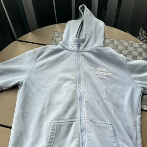 Ljusblå jacka från Peak Performance - En ljusblå jacka från Peak Performance med dragkedja och huva. Jackan har två framfickor och ribbstickade muddar vid ärmslut och nederkant. Märkets logotyp är broderad på bröstet. Säljer pga att den är för liten, passformen är bra. Ny pris 800kr, priset kan diskuteras