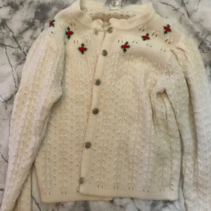 Vit stickad kofta med broderade blommor - En vit stickad kofta med broderade röda och gröna blommor vid axlarna. Koftan har långa ärmar och knappar framtill. Den är varm och bekväm.Inte bästa bilderna men skriv om ni behöver mer