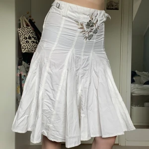 Vit bomullskjol med broderier - Midikjol med gröna, silvriga och ljusbruna blommiga broderier, ficka på baksidan och små spännen i båda sidorna. 