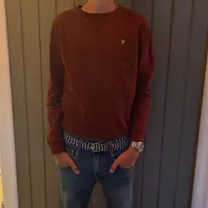 En stilren vinröd tröja från Lyle & Scott Junior. Tröjan har en rund halsringning och ett broderat gult logomärke på bröstet. Den är långärmad och tillverkad i ett mjukt material.