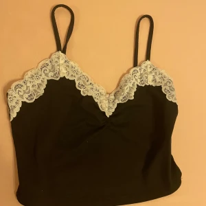 Svart linne med spetsdetaljer - Ett svart linne med smala axelband och vita spetsdetaljer längs halsringningen och ärmhålen. Linnet har en enkel och elegant design. Aldrig använt. Skriv till mig om ni har frågor. Pris kan bli sänkt 💕 