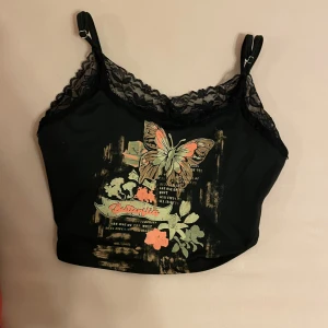 Svart linne med fjärilsmotiv och spetsdetaljer - Ett svart linne med justerbara axelband och spetsdetaljer vid halsringningen. Linnet har ett färgglatt fjärilsmotiv och blommor på framsidan. Skriv gärna om ni har frågor 💕