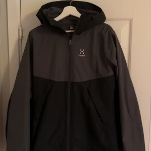 Haglöfs Jacka - Gråsvart Haglöfs jacka som är vind- och vattentät. Inga fläckar eller hål. Storlek S men är väldigt stor i storleken, passar mig som vanligtvis har storlek L och är 190cm, ca 70kg. Finns spännen så man kan justera passform. Inget kvitto kvar tyvärr.