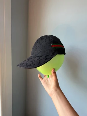 Svart keps med GG-mönster och ränder - Snygg svart keps.På sidan finns en grön och röd rand som ger en ikonisk touch. Kepsen har böjd skärm och är tillverkad i tygmaterial. Perfekt för dig som vill ha en stilren accessoar.