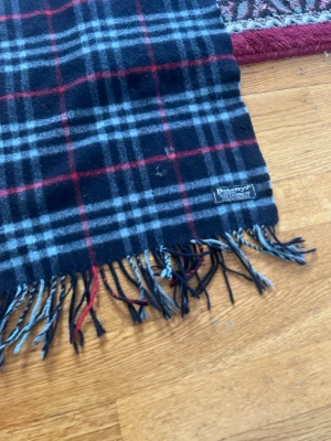 Rutig halsduk i ull från Burberry - Klassisk halsduk från Burberry i svart, grått och rött rutmönster. Tillverkad i mjuk ull med fransar i ändarna. Perfekt accessoar för att lyfta din outfit med en tidlös och elegant touch.