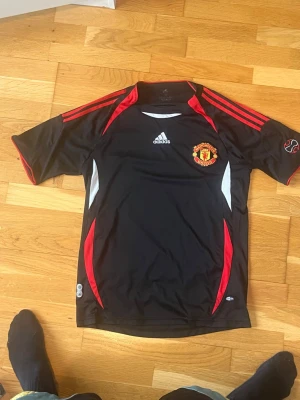 Manchester United svart tränings t-shirt - Snygg svart tränings t-shirt från Adidas med Manchester Uniteds klubbmärke på bröstet. Tröjan har röda och vita detaljer, korta ärmar och klassiska Adidas-ränder på axlarna. Perfekt för fotbollsträning eller att bära till vardags.