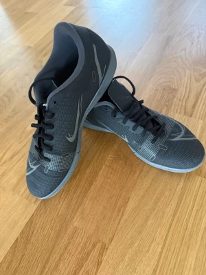 Svarta Nike Mercurial fotbollsskor - Säljer ett par svarta Nike Mercurial fotbollsskor med gråa detaljer och snörning. Skorna har en låg profil och är tillverkade i syntetmaterial för bra bollkontroll. Snygg design med grafiska mönster på ovandelen och klassisk Nike-logga på sidan.