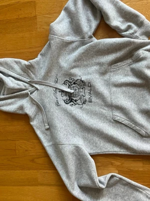 Grå hoodie med tryck från Bvaldi - Snygg grå hoodie från Bvaldi med stort svart tryck på bröstet och texten 'Cote d'Azur Club'. Hoodien har klassisk känguruficka och snörning i huvan. Perfekt för chill dagar eller när du vill ha en clean streetstyle-look.