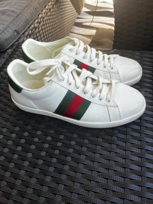 Vita sneakers med gröna och röda detaljer - Snygga vita sneakers med gröna och röda ränder på sidan samt grön mönstrad detalj på hälen. Skorna har klassisk låg profil, vita snören och är tillverkade i slätt läder. Perfekta för dig som vill ha en clean och stilren look.