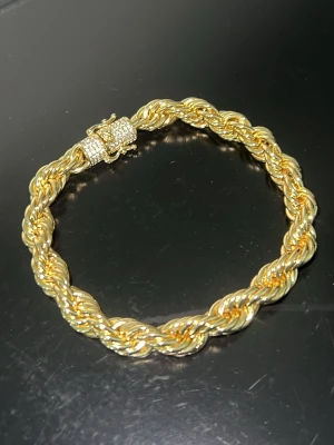 Cordell Armband 8mm  - Säljer ett snyggt armband i guldfärg med tvinnad repdesign. Låset är dekorerat med små glittrande stenar som ger extra bling. Perfekt accessoar för dig som gillar statement-smycken och vill sticka ut lite extra.