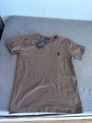 Brun t-shirt från Polo Ralph Lauren - Snygg brun t-shirt från Polo Ralph Lauren med klassisk passform och korta ärmar. T-shirten har en liten mörkblå logga broderad på bröstet och är gjord i mjuk bomull. Perfekt till jeans eller shorts för en clean look.