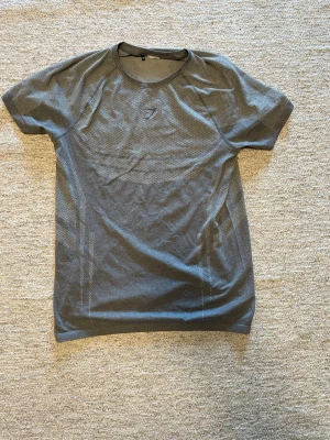 Grå träningströja från Gymshark - Snygg grå träningströja från Gymshark med korta ärmar och rund hals. Tröjan har ett diskret Gymshark-logotyp på bröstet och är tillverkad i ett lätt, ventilerande material som passar perfekt för gymmet eller löprundan.