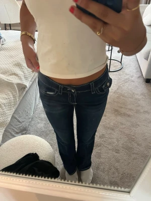 Mörkblå low waist jeans med kontrastsömmar - Säljer ett par mörkblå low waist jeans med raka ben och tydliga vita kontrastsömmar. Jeansen har stora fickor både fram och bak samt dekorativa knappar på bakfickorna. Perfekta för en trendig och avslappnad look.