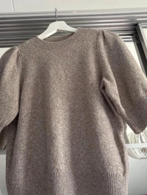 Beige stickad tröja Vero Moda - Mysig beige stickad tröja från Vero Moda med puffärmar och ribbade muddar vid hals, ärmslut och nederkant. Tröjan har en avslappnad passform och är perfekt för kyliga dagar. Materialet är mjukt och ger en skön känsla.