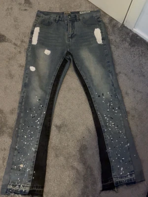 Grå jeans med färgstänk och slitningar - Unika grå jeans med svarta paneler längs benen, vita färgstänk och slitna detaljer framtill och baktill. Raka ben med råa kanter nertill ger en edgy look. Perfekta för dig som vill sticka ut med din stil. Dessa är gallery dept 