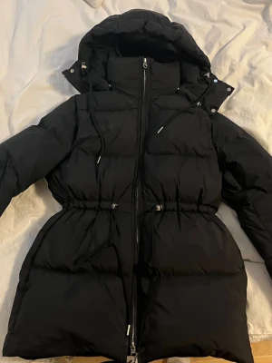 Svart dunjacka med huva - Snygg svart dunjacka med stor huva och dragsko  i midjan. Jackan har tvåvägsdragkedja framtill och justerbara detaljer vid huvan. Använd två gånger. Mycket bra skick, storlek XS. Köpt för 2290 kr hos arket!