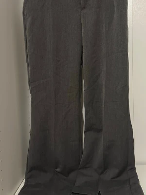 Mörkgrå vida kostymbyxor - Snygga mörkgrå kostymbyxor med utsvängda ben och klassisk passform. Byxorna har en stilren look och är perfekta för dig som gillar en trendig och clean stil. Materialet ser ut att vara en mjuk och lätt vävd textil som ger en dressad känsla.