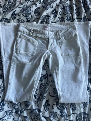 Vita bootcut jeans med fickor - Säljer ett par vita jeans med utsvängda ben och två stora fickor framtill. Jeansen har hög midja med dubbla knappar och bälteshällor. Materialet är denim och de har slitningar vid knäna för en trendig look.