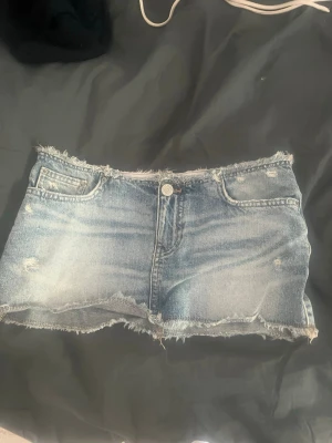 Blå jeanskjol  - Säljer en kort jeanskjol i blå denim med slitna detaljer och råa kanter. Kjolen har klassiska fickor fram och knappstängning. Perfekt för dig som gillar en avslappnad och trendig stil.